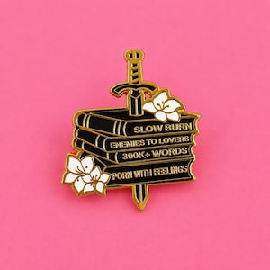 May include: A gold-toned metal enamel pin featuring a stack of black books pierced by a sword. The books are inscribed with the text SLOW BURN, ENEMIES TO LOVERS, 300K+ WORDS, and PORN WITH FEELINGS. White floral accents decorate the corners.