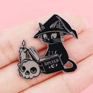 Peut inclure: Une broche en émail noir représentant un chat portant un chapeau de sorcière pointu, assis à côté d'un livre ouvert marqué SPELLS avec un symbole de croissant de lune, une bougie allumée et un petit crâne. Le design utilise des contours en métal.