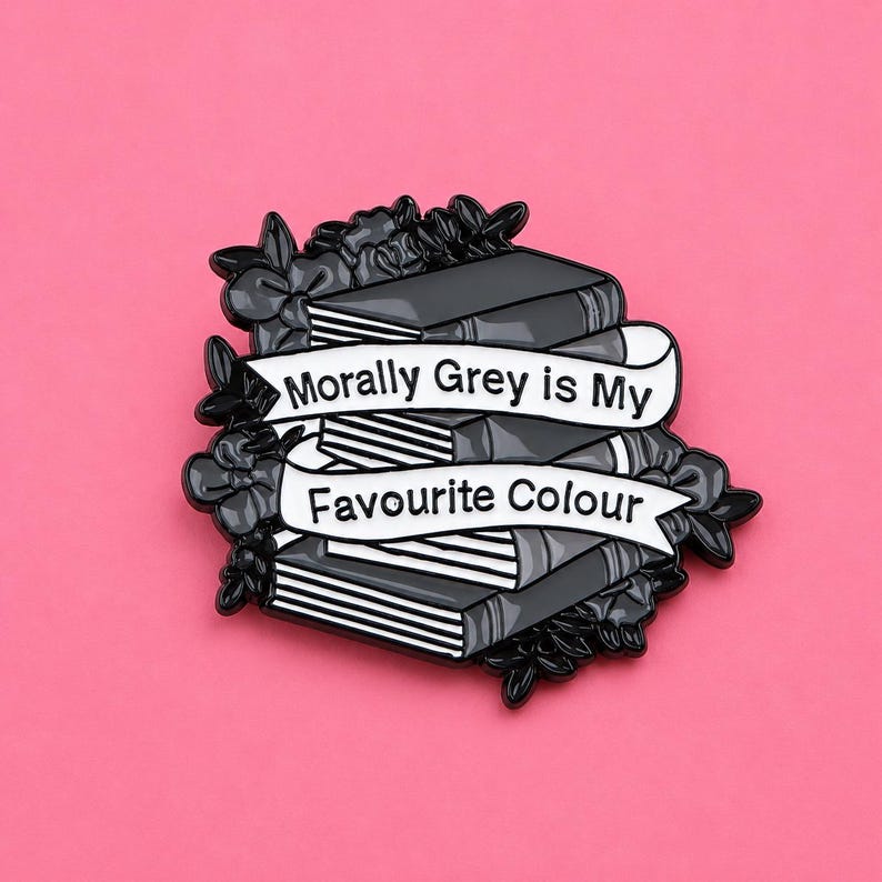 May include: A black and white enamel pin featuring a stack of three books surrounded by dark floral elements. A white banner draped across the books displays the black text, Morally Grey is My Favourite Colour, in a clear, sans-serif font.