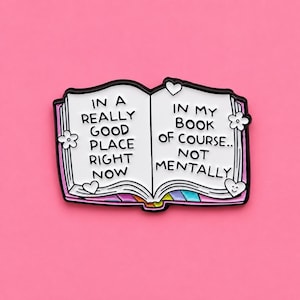Peut inclure: Une broche en émail en forme de livre ouvert avec des pages blanches et un texte noir indiquant : IN A REALLY GOOD PLACE RIGHT NOW IN MY BOOK OF COURSE.. NOT MENTALLY. La broche présente de petites fleurs blanches, des cœurs et des bords arc-en-ciel.