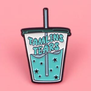 May include: A teal and white enamel pin in the shape of a beverage cup. The pin features the text "TAMLINS TEARS" in teal, with teal liquid and black star accents. The pin has a teal straw and a black outline.