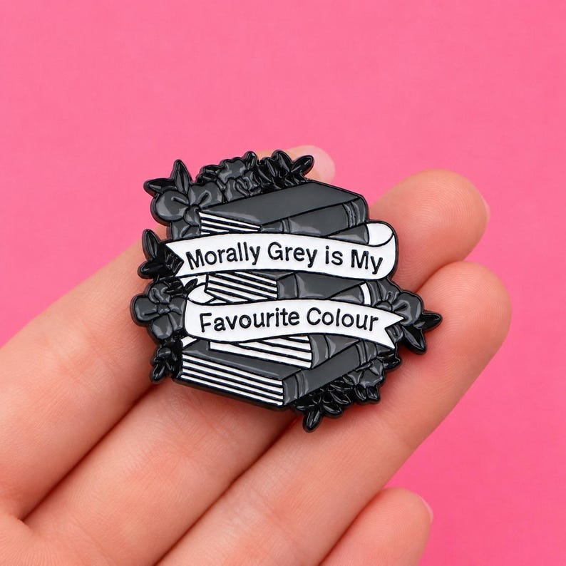 May include: A black and white enamel pin featuring a stack of books surrounded by floral elements. A white banner draped across the books contains the black text "Morally Grey is My Favourite Colour" in a clear, sans-serif font style.
