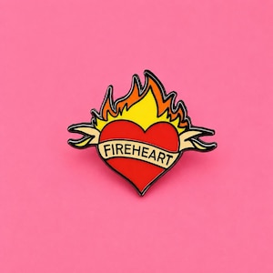 May include: A metal enamel pin shaped like a red heart with yellow and orange flames rising from the top. A cream-coloured banner wraps across the centre of the heart with the word FIREHEART printed in black capital letters. The design has a black outline.
