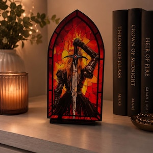 Peut inclure: Une lampe en forme d'arche style vitrail représentant une silhouette en armure sombre tenant une épée dressée devant un fond de flammes orange et rouges vives. La lampe possède un cadre noir et est placée entre trois livres.