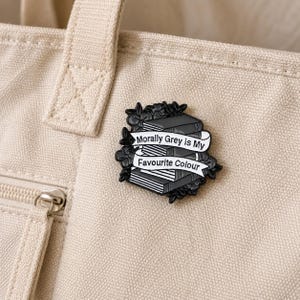 May include: A black and white enamel pin featuring a stack of books surrounded by floral details. A white banner across the center displays the text Morally Grey is My Favourite Colour in black lettering, with a dark outline and metallic finish.