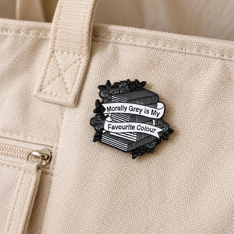May include: A black and white enamel pin featuring a stack of books surrounded by floral details. A white banner across the center displays the text Morally Grey is My Favourite Colour in black lettering, with a dark outline and metallic finish.