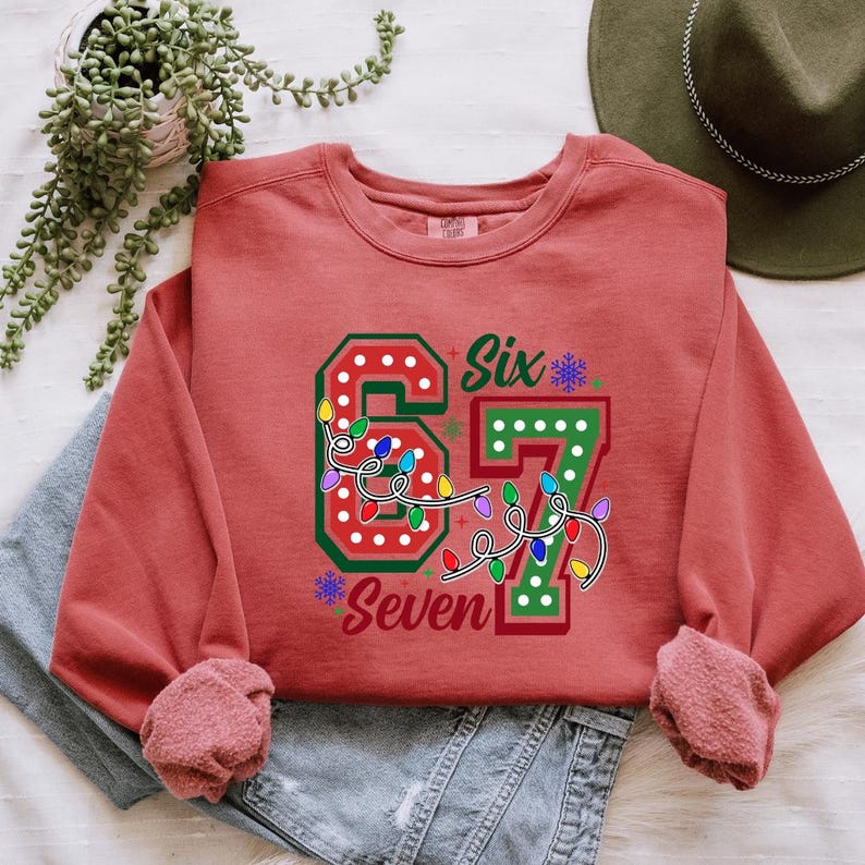 A red sweater with the number "67" and the word "Seventh" printed on it, along with a hat and some plants, is laid out on a surface.