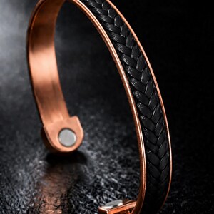 Op de afbeelding: Een koperkleurige armband met een centrale zwarte gevlochten inleg. De armband heeft een gladde metalen afwerking aan de buitenranden en bevat twee kleine ronde zilverkleurige magneten aan de open uiteinden van de band.