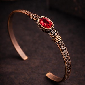 Könnte beinhalten: Ein bronzefarbenes Armreif-Armband mit einem ovalen, roten Edelstein in der Mitte. Das Armband weist aufwendige Spiral- und geätzte Designs auf. Die offenen Enden ermöglichen ein einfaches Tragen. Der Schmuck ist vor einem dunklen Hintergrund platziert.