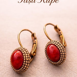 May include: Two gold-coloured earrings, each featuring a red, oval-shaped stone. The stones are framed by a textured, grid-like border. The earrings have a lever-back clasp. The text "Kırmızı Akik Taşlı Küpe" is visible in the image.
