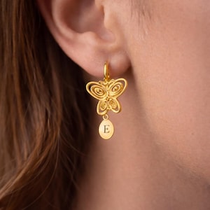 Può includere: Orecchini a farfalla color oro con chiusura a cerchio. Il design a farfalla presenta intricati dettagli traforati. Un piccolo ciondolo ovale con la lettera "E" pende sotto. Gli orecchini sono indossati.