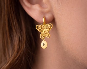 May include: Gold-colored butterfly earrings with a hoop closure. The butterfly design features intricate cut-out details. A small, oval charm with the letter "E" dangles below the butterfly. The earrings are shown on a person's ear.