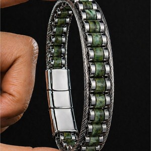 Könnte beinhalten: Ein Armband mit einer zentralen Reihe aus zylindrischen, grün gesprenkelten Steinperlen, die von zwei silberfarbenen, geflochtenen Metallkabeln eingerahmt werden. Das Design umfasst einen polierten, rechteckigen Magnetverschluss aus Metall.
