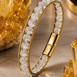 May include: A bracelet featuring a row of clear, spherical beads flanked by two thin, braided gold-tone metal wires. The design includes a polished gold-tone cylindrical magnetic clasp that secures the two ends of the bracelet together.