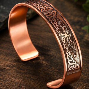 May include: A copper cuff bracelet featuring an embossed design of a tree with intricate, interwoven Celtic knotwork patterns. The metal has a polished finish on the edges and a darkened, recessed texture within the detailed engraved artwork.