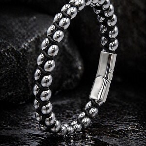 Puede incluir: Una pulsera que presenta dos filas paralelas de cuentas esféricas de color plateado pulido, entrelazadas con un cordón negro. El diseño incluye un cierre metálico rectangular y liso de color plateado que une los extremos de las hebras.