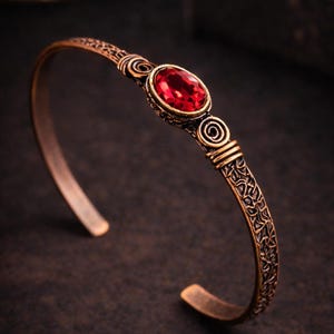 May include: A copper-toned metal cuff bracelet featuring an oval-shaped, faceted red gemstone at the centre. The band displays intricate, raised scrollwork and spiral patterns along its length, with a dark, weathered finish throughout the metal.