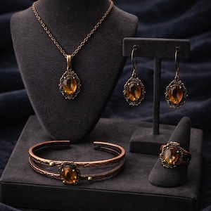 Può includere: Un set di gioielli composto da collana, orecchini, bracciale e anello. Ogni pezzo presenta una pietra ovale color ambra incastonata in un metallo color bronzo con dettagli ornati. I gioielli sono esposti su supporti in velluto grigio scuro su sfondo blu scuro.