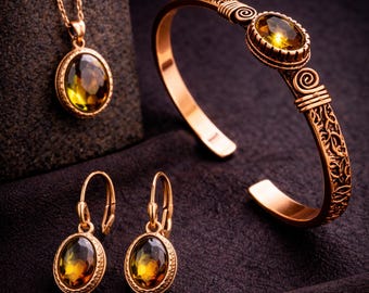 May include: A gold-toned jewelry set featuring a necklace, earrings, and a bracelet. Each piece has an oval-shaped, amber-colored stone. The bracelet has an open design with decorative scrollwork. The jewelry is displayed on a dark, textured surface.