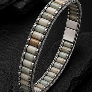 May include: A bracelet featuring a series of cylindrical, multi-colored stone beads in shades of cream, beige, and pale green, set within a silver-toned metal frame. The design includes a woven silver-toned mesh band and a rectangular metal clasp.