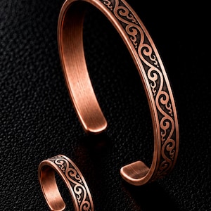 May include: A copper-toned metal cuff bracelet and a matching open ring, both featuring a dark, recessed swirling scroll pattern engraved along the outer surface. The metal has a brushed finish with polished edges, showcasing a continuous design.