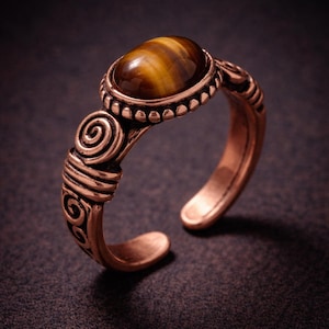 May include: A bronze-colored adjustable ring featuring an oval tiger's eye stone. The ring has an open design with decorative spiral and line details on the band. The stone is a warm brown color with golden stripes.