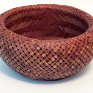 May include: A woven basket made of natural materials, with a reddish-brown color and a round shape. The basket is made with a tight weave and has a smooth, polished finish.