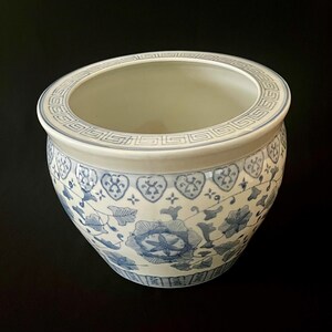 May include: A white ceramic planter featuring a blue floral pattern and a Greek key design around the rim. The bowl-shaped vessel has a smooth, glossy finish with intricate blue illustrations of flowers and leaves covering the exterior surface.