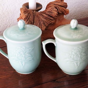 May include: Two light green ceramic teacups with lids. The cups have a floral design and are on a wooden surface.