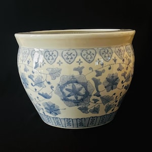 May include: A round ceramic planter featuring a cream-colored base with intricate blue floral patterns and vine motifs. The upper rim has a decorative border of heart-shaped designs, while the base features a repeating vertical line pattern.