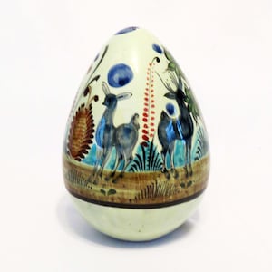 May include: A white ceramic egg-shaped ornament with a hand-painted design of deer and foliage in shades of blue, green, red, and brown.
