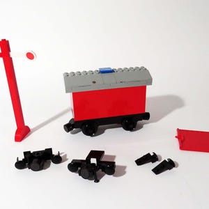 May include: A red Lego train car with a gray roof and a blue detail. The car is on a white surface with a red Lego signal and black Lego wheels.
