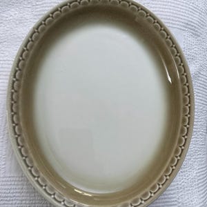 May include: Oval-shaped ceramic serving dish with a scalloped edge. The dish features a gradient of colors, transitioning from a creamy white center to a light brown border. The rim has a repeating pattern of small, rounded shapes.