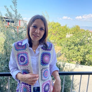 Puede incluir: Un chaleco de ganchillo sin mangas con un patrón de cuadrados de la abuela multicolores en tonos morado, rosa y azul, unidos por un borde color crema. El chaleco se lleva sobre una camisa blanca de botones y pantalones vaqueros azules.