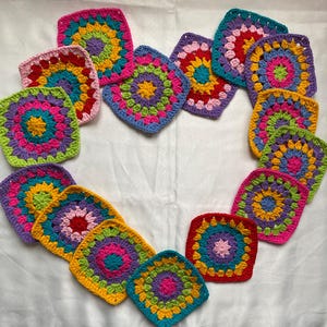 May include: A collection of colorful, crocheted squares arranged in a heart shape. Each square features a vibrant, concentric circle design with shades of pink, green, yellow, blue, and purple. The squares are set against a white background.