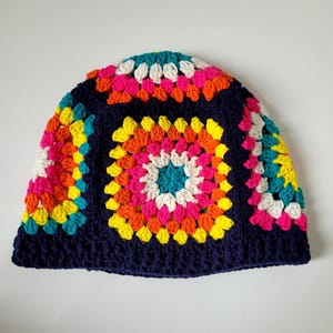Puede incluir: Un sombrero de pescador de ganchillo construido a partir de varios cuadrados de la abuela. El sombrero presenta un color base azul marino oscuro con vibrantes acentos de hilo multicolor en tonos rosa brillante, naranja, amarillo, blanco y verde azulado.