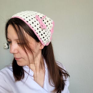 May include: A white crochet headscarf with a pink border and a pink bow. The head covering is made with an open weave pattern. The headscarf is worn on the head.