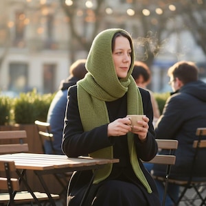 May include: A woman wearing a green knit hood and scarf, holding a beige mug. She is seated at a wooden table outdoors, with other people and a blurred background. The scarf and hood are a solid, light green color.