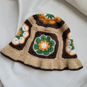 May include: A crochet bucket hat featuring a patchwork design of floral granny squares in shades of dark brown, tan, forest green, white, and golden yellow. The hat has a wide, slightly flared brim and a rounded crown constructed from yarn.
