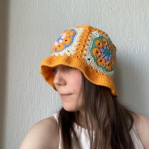 May include: A crochet bucket hat featuring a vibrant orange base color with multi-colored granny square motifs. The hat displays intricate floral patterns in shades of light blue, lavender, and cream, constructed with a textured yarn stitch.