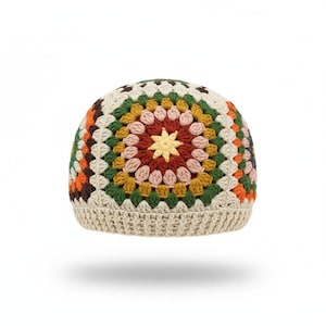 Op de afbeelding: Een gehaakte muts met een veelkleurig granny square patroon. Het ontwerp bevat concentrische cirkels van crème, donkergroen, mosterdgeel, oudroze en donkerrood garen, afgewerkt met een effen crème-kleurige geribbelde boord.