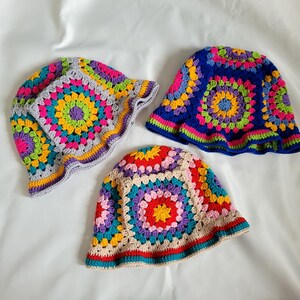 May include: Three crochet bucket hats featuring colorful granny square patterns. One hat has a light gray base, another has a deep blue base, and the third has a beige base. Each hat displays vibrant circular motifs in yellow, pink, purple, and teal.