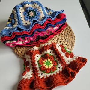 Peut inclure: Plusieurs chapeaux seau crochetés à la main, aux couleurs vives. Les chapeaux présentent un motif carré dans des tons de bleu, de rouge et d'orange. Les chapeaux ont un bord à volants et sont présentés sur un tapis tissé.