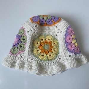 May include: A crochet bucket hat featuring a white base with multi-colored granny square motifs. The circular patterns incorporate shades of yellow, orange, sage green, lavender, and soft pink, creating a vibrant, textured, and handcrafted appearance.