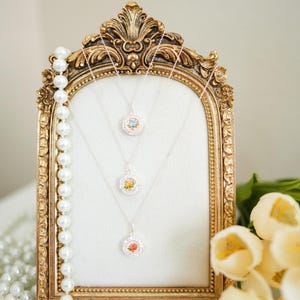 May include: Three silver necklaces with floral pendants are displayed on a gold ornate frame. A strand of pearls hangs on the left. White tulips are on the right. The pendants have colorful flower designs.