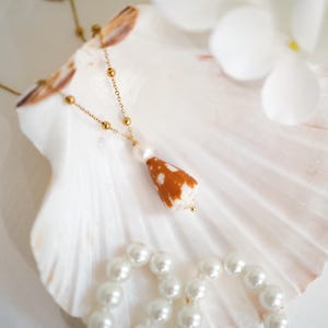 Puede incluir: Un collar dorado con un colgante de concha y una perla. El collar descansa sobre una concha grande y blanca, con un collar de perlas de imitación y flores blancas en el fondo. El colgante es marrón y blanco.