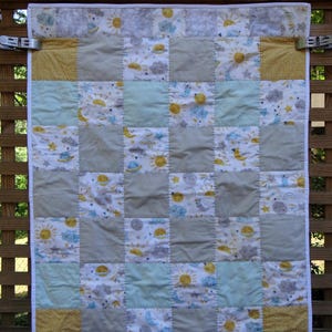 May include: A patchwork quilt with a celestial theme, featuring squares of light blue, gray, and yellow fabrics. The quilt showcases suns, moons, clouds, and stars. The quilt is hanging on a wooden fence.