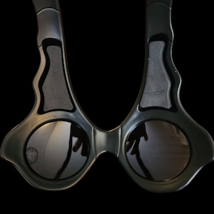 May include: Black sunglasses with a unique, angular design. The frames are dark gray with a matte finish, and the lenses are round and reflective. The arms curve inward, and the overall shape is modern and stylish.