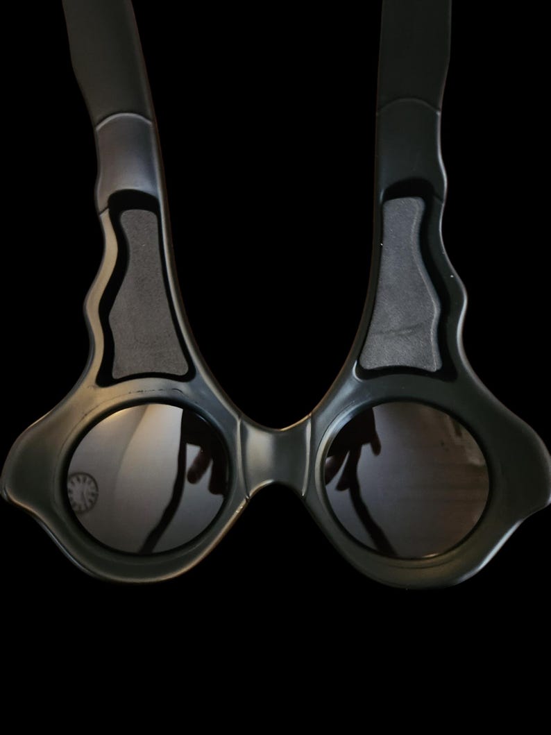 May include: Black sunglasses with a unique, angular design. The frames are dark gray with a matte finish, and the lenses are round and reflective. The arms curve inward, and the overall shape is modern and stylish.