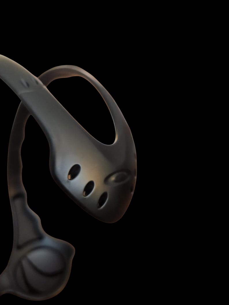 May include: A dark gray kitchen ladle with a curved handle and a rounded head featuring three oval holes. The ladle is made of a smooth, possibly metallic material, and is set against a black background.